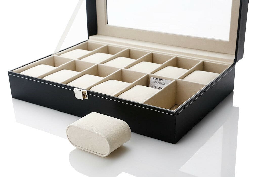 Coffret à montres noir avec couvercle en verre, contenant seize compartiments beiges. Un coussin de compartiment est placé devant le coffret. Image